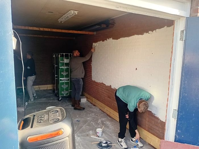 Drie mensen schilderen de bakstenen muur van een ruimte wit.