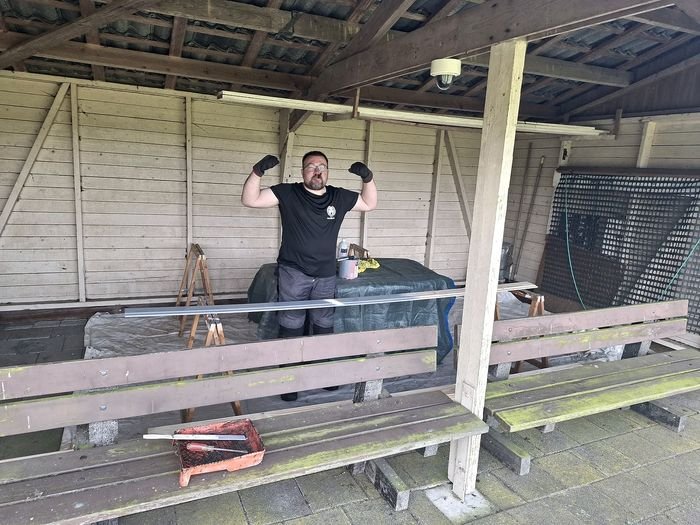 Een man in werkhandschoenen steekt zijn armen in de lucht onder een houten overkapping vol bankjes.