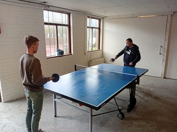 Twee mensen spelen tafeltennis in een lichte ruimte met grote ramen.