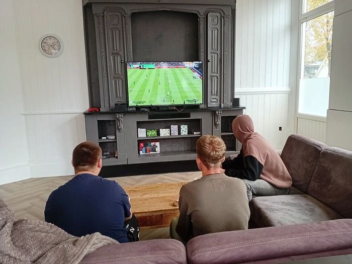 Drie bewoners zitten op de bank in de woonkamer en kijken voetbal op tv.