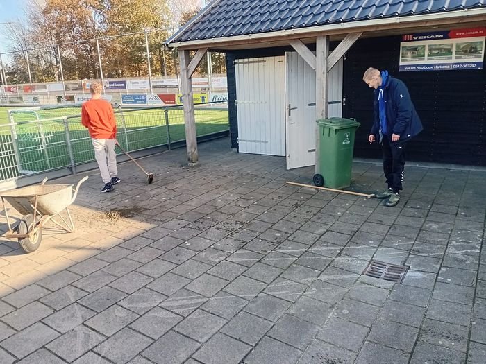 Twee mensen vegen het terras naast een houten overkapping langs het voetbalveld.