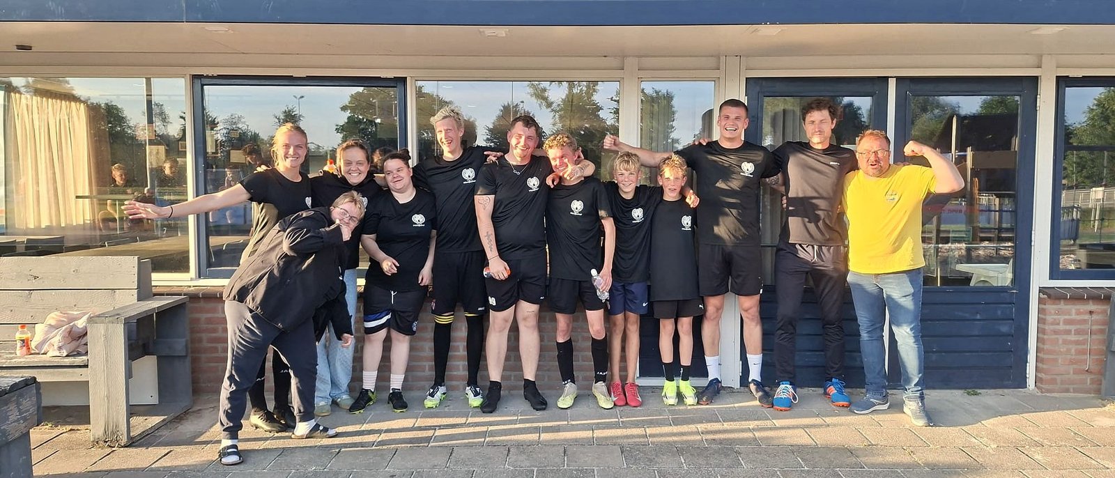 Een groep bewoners en begeleiders in zwarte shirts staat lachend voor een sportkantine.