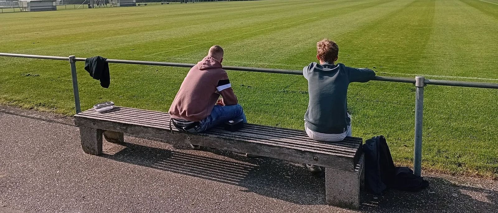 Twee bewoners zitten op een bankje langs een voetbalveld en kijken uit over het gras.
