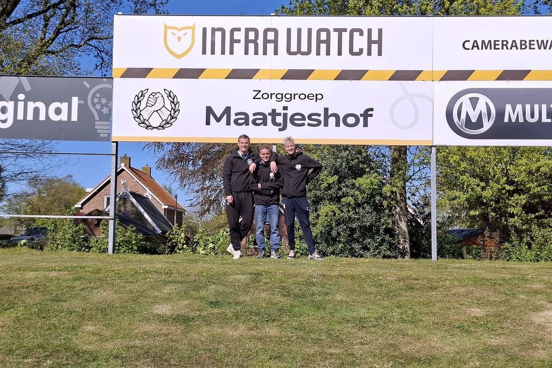 Bewoners en een begeleider staan op het veld onder een reclamebord van Zorggroep Maatjeshof.