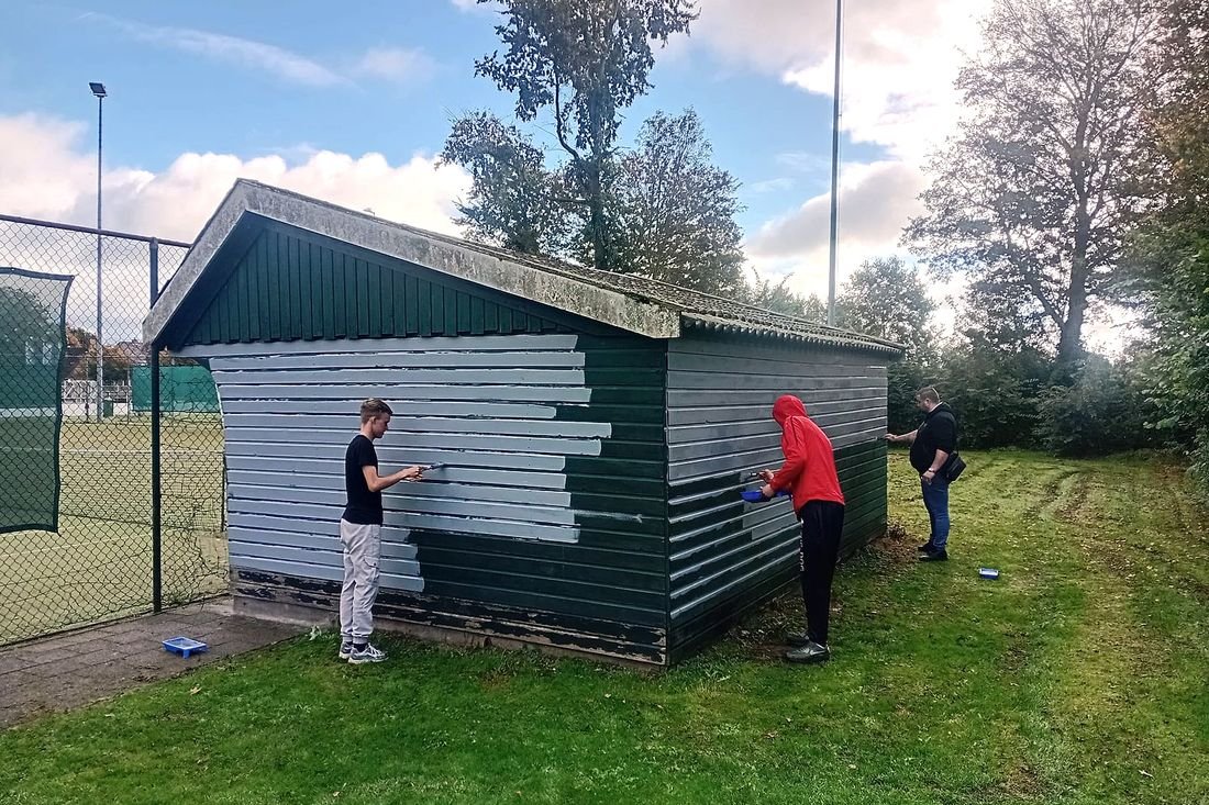 Drie bewoners schilderen een groene houten schuur langs het voetbalveld in een lichte kleur.